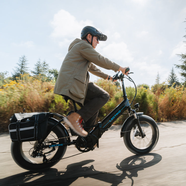 Man wearing a helmet is riding a Lectric XPremium eBike with water-resistant pannier bags in motion on a bright sunny road
