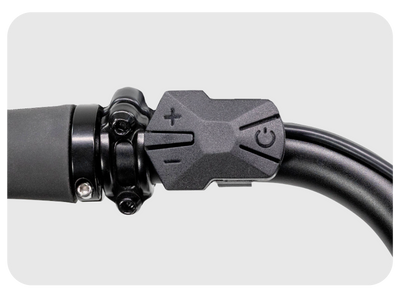 Close-up of a black handlebar-mounted control switch on the XP 3.0 Long-Range eBike. The switch features a power button icon, a plus sign, and a minus sign, suggesting it controls power or speed settings. The metallic handlebar and control switch are set against a plain gray background, ready for your next adventure.