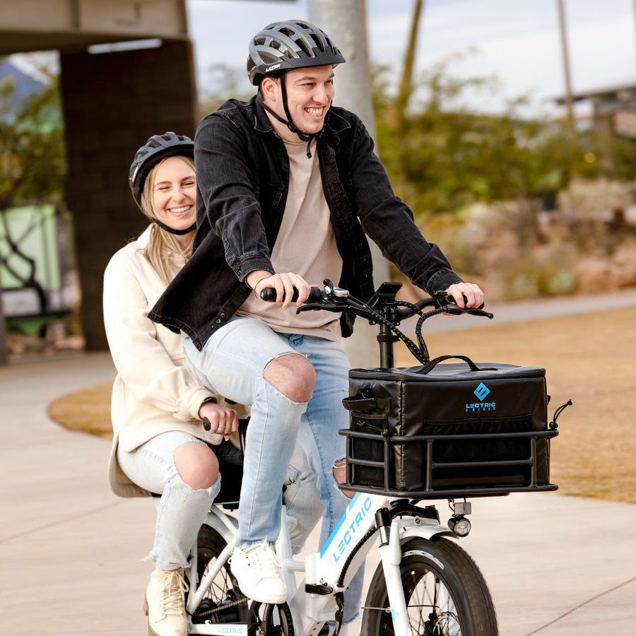 Man and woman smiling while riding an ebike through the park with a soft cooler in the front basket